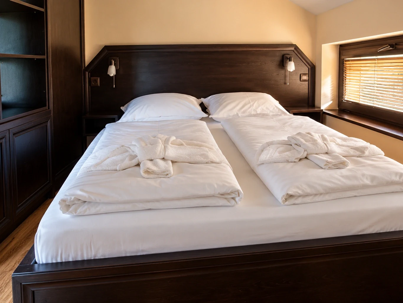 The double bed with white linen and folded towels beside the window with blinds
