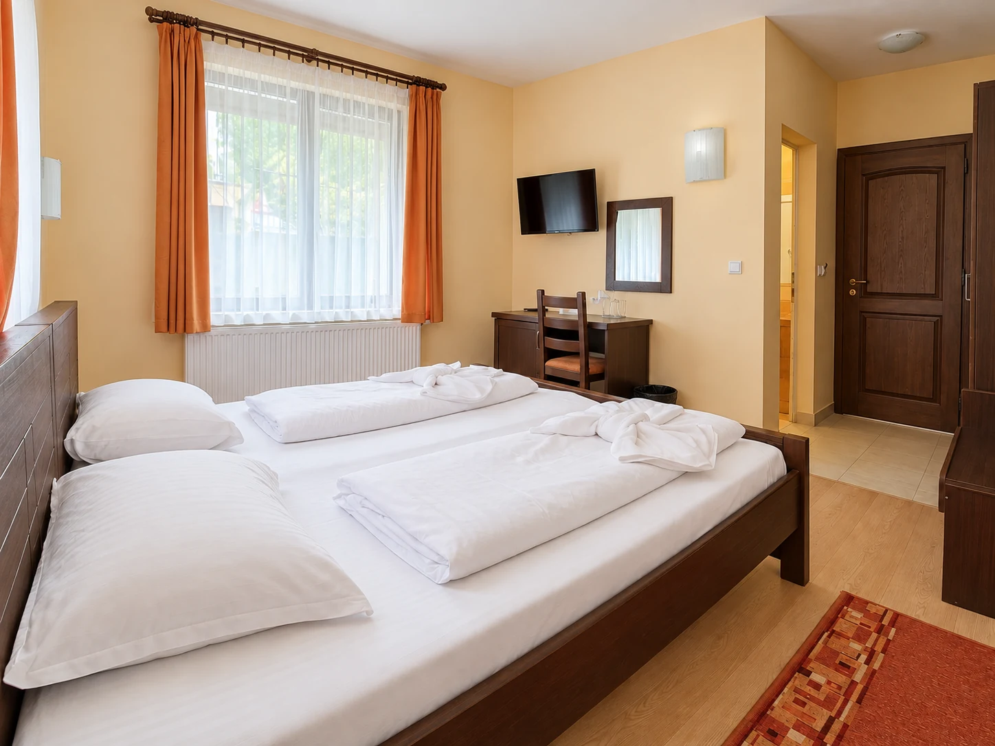 A wide bed with a headboard of dark wooden boards, white bedding, a desk and a window with orange curtains