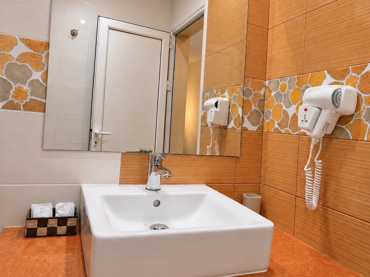 A white basin on an orange counter, a mirror, a hairdryer and a band of pebble-pattern tiles