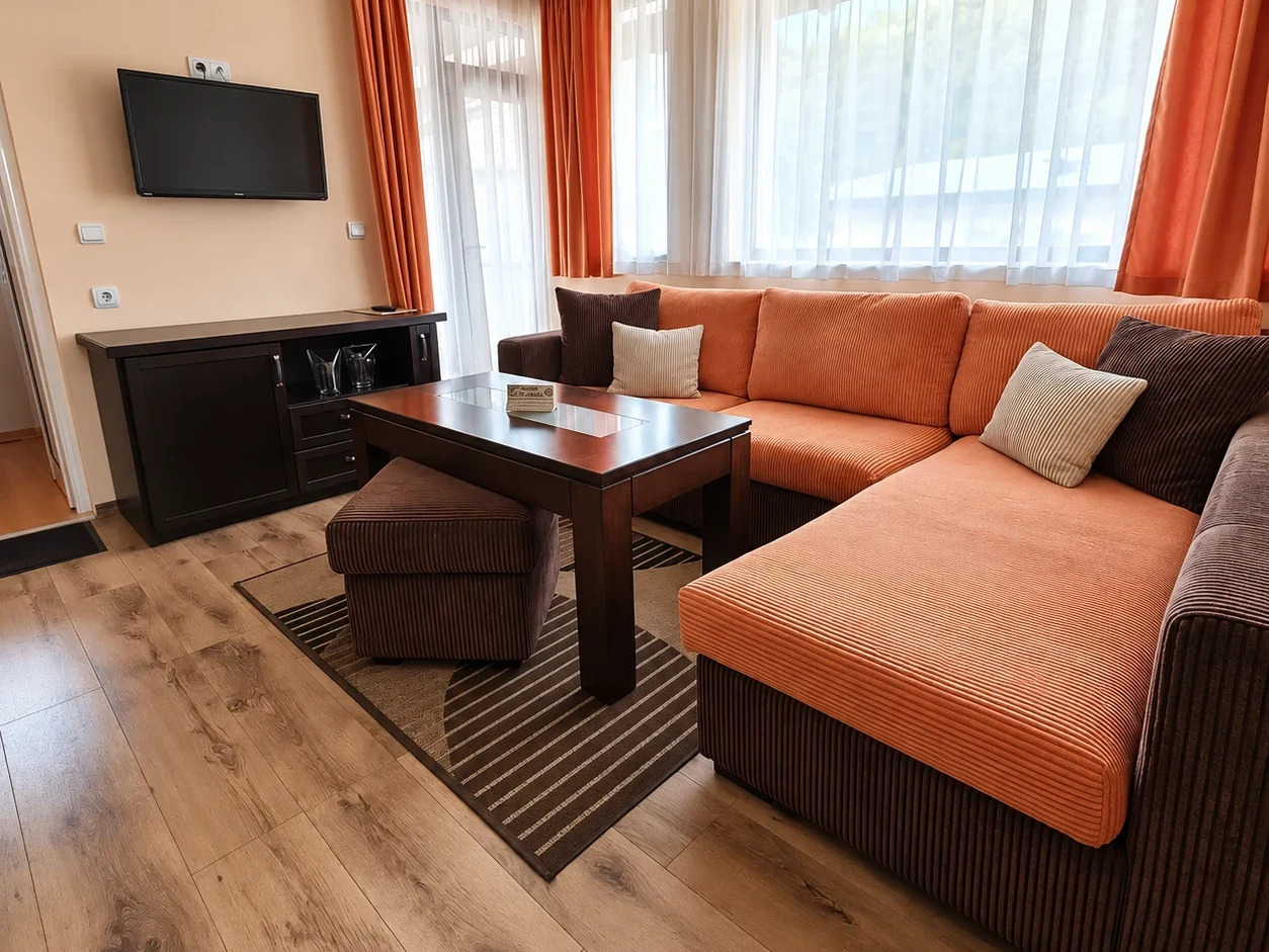 A living room with an orange corner sofa, a footstool, a low wooden table and a large window with sheer and orange curtains