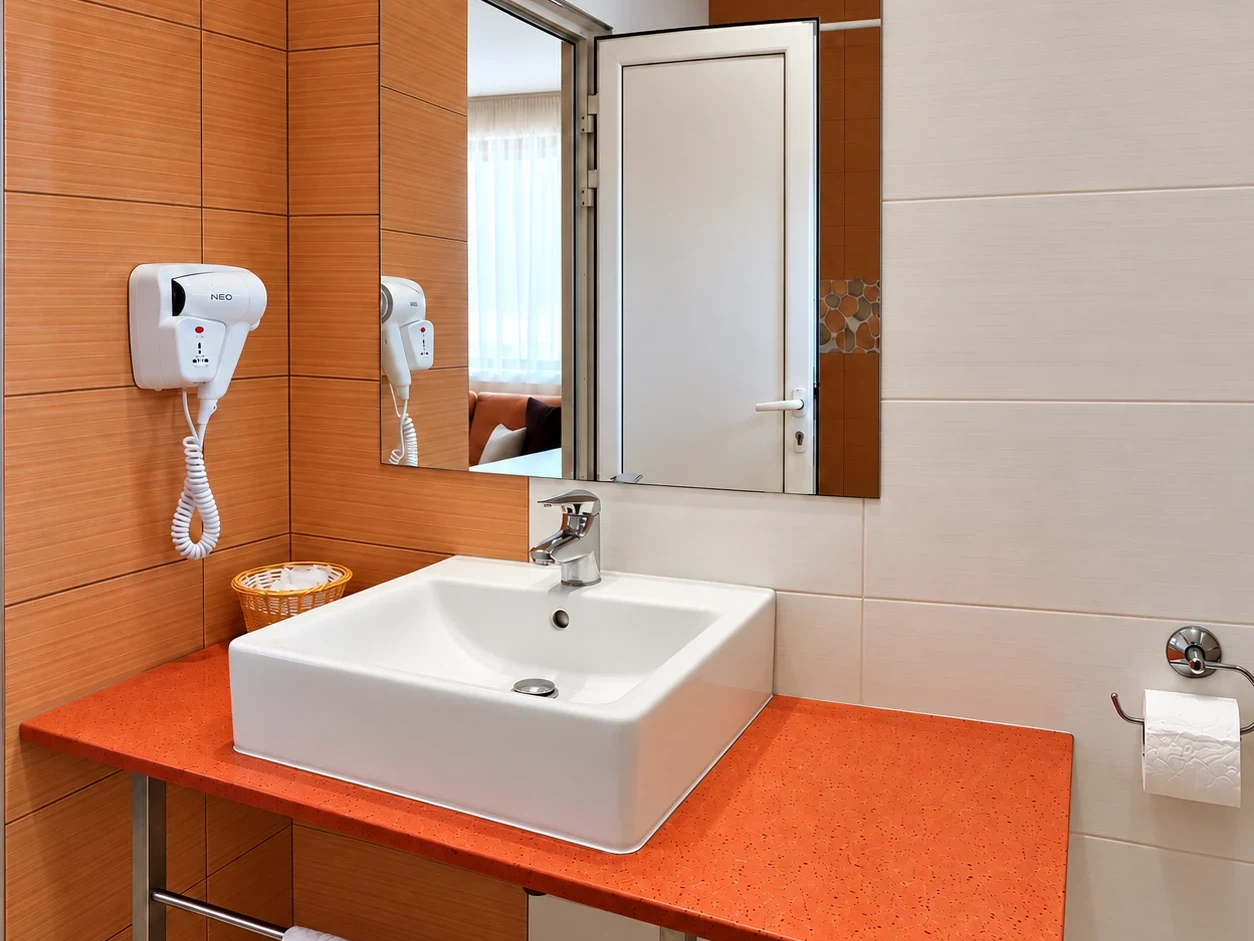 A white basin on an orange counter, a mirror, a hairdryer and white and orange tiles
