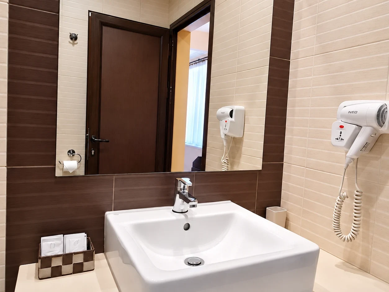 A white basin on a pale counter, a wide mirror, a hairdryer and a wall of dark brown tiles