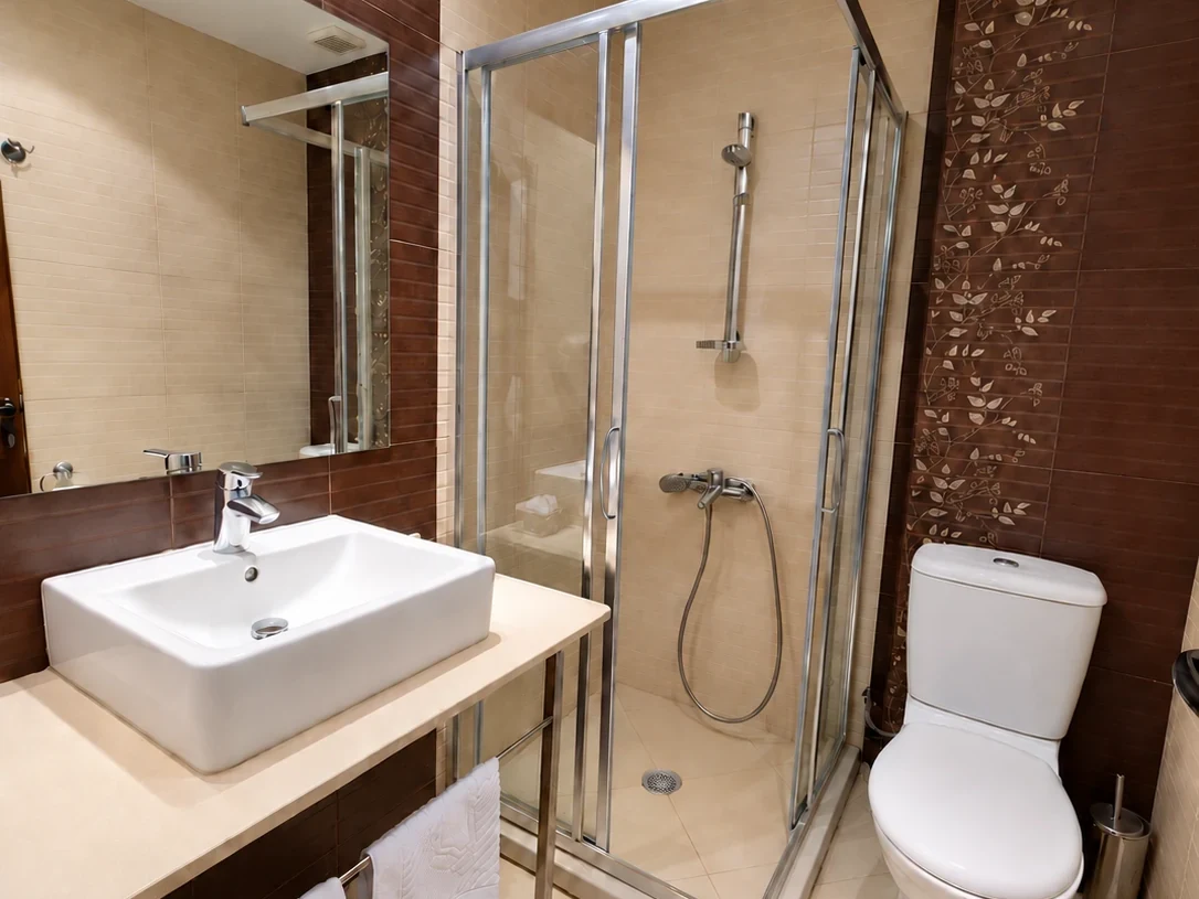 A basin on a pale counter, a shower cubicle, a toilet and a wall of dark floral-motif tiles