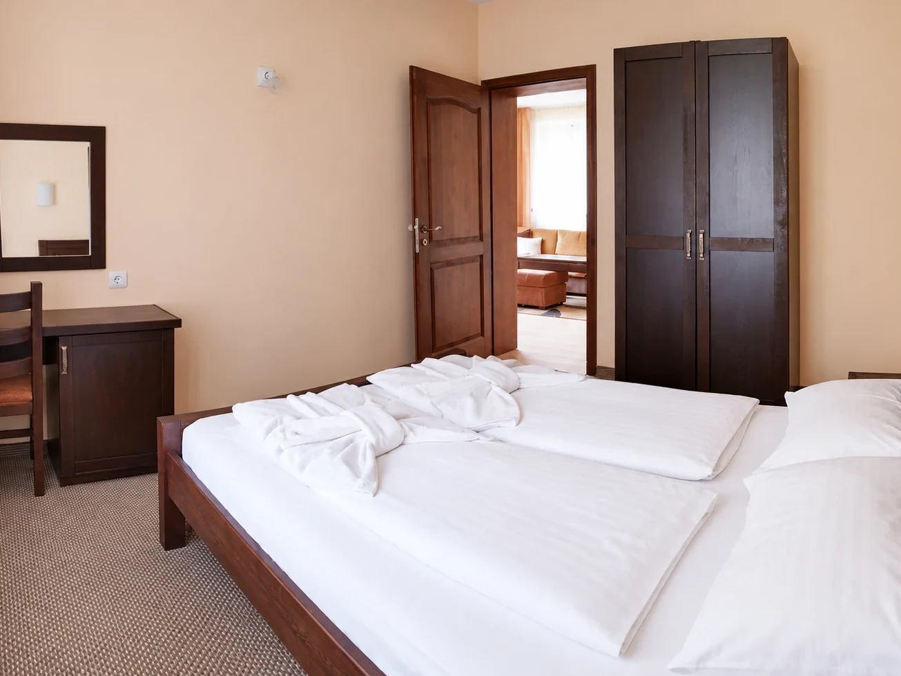 A double bed with white bedding, a desk and chair, a mirror, a dark wardrobe and an open door