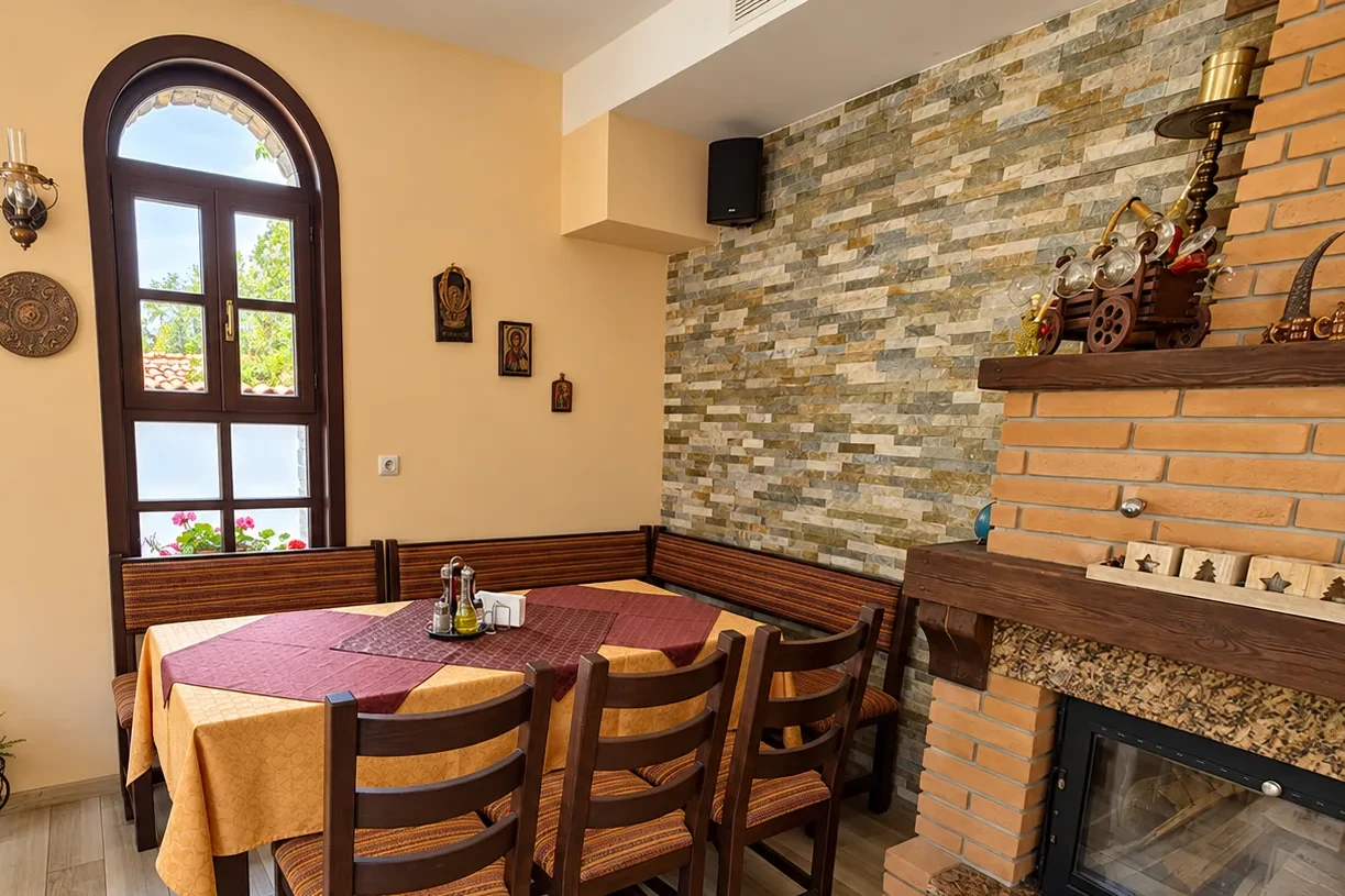 A corner of the restaurant with a table for six, upholstered benches, arched windows with pot plants and a brick fireplace